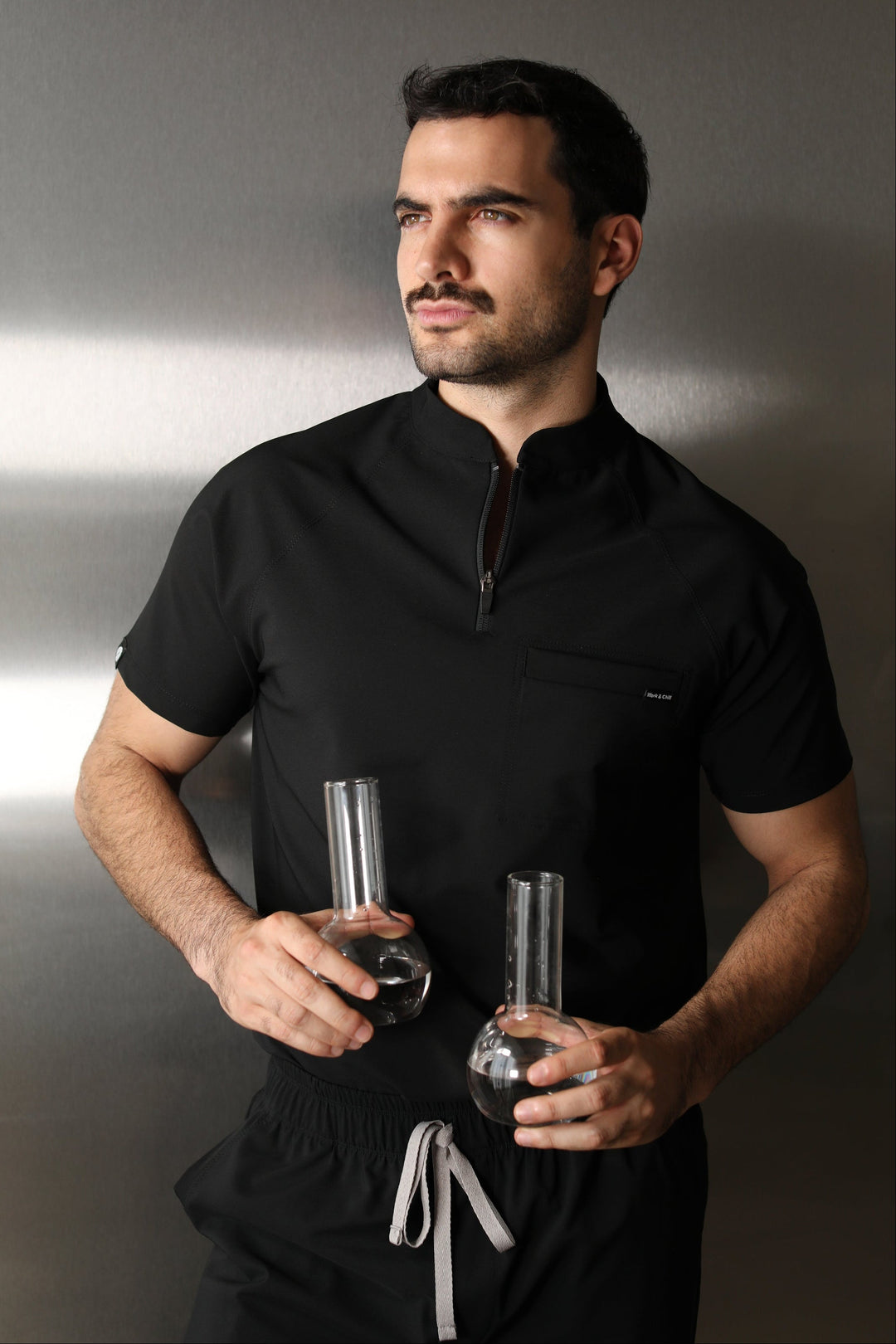 Man wearing the Harry Black W&amp;C Shield™ scrub top while holding two laboratory flasks filled with clear liquid.
